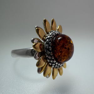 May include: A silver ring with a sunflower design. It has gold-coloured petals, a silver band, and a large, round, amber-coloured stone at the centre. This ring is a piece of jewellery.