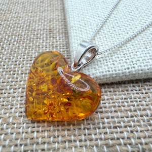 Op de afbeelding: Een hartvormige barnsteen hanger met een zilveren balein en ketting. De barnsteen heeft een warme, doorschijnende oranje kleur met zichtbare insluitsels. De hanger ligt op een getextureerd, neutraal getint oppervlak.