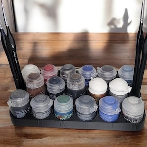May include: A black plastic paint pot holder with 18 small paint pots, some with lids, arranged in rows. The pots are filled with various colors of paint, including black, white, blue, green, and brown. Two paint brushes are on the left and right sides of the holder.