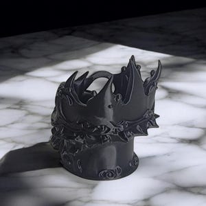 K&ouml;nnte beinhalten: Ein schwarzer, kronenf&ouml;rmiger Beh&auml;lter. Der Beh&auml;lter hat ein detailliertes Design mit scharfen Spitzen und eine strukturierte Oberfl&auml;che. Er steht auf einer Marmoroberfl&auml;che.