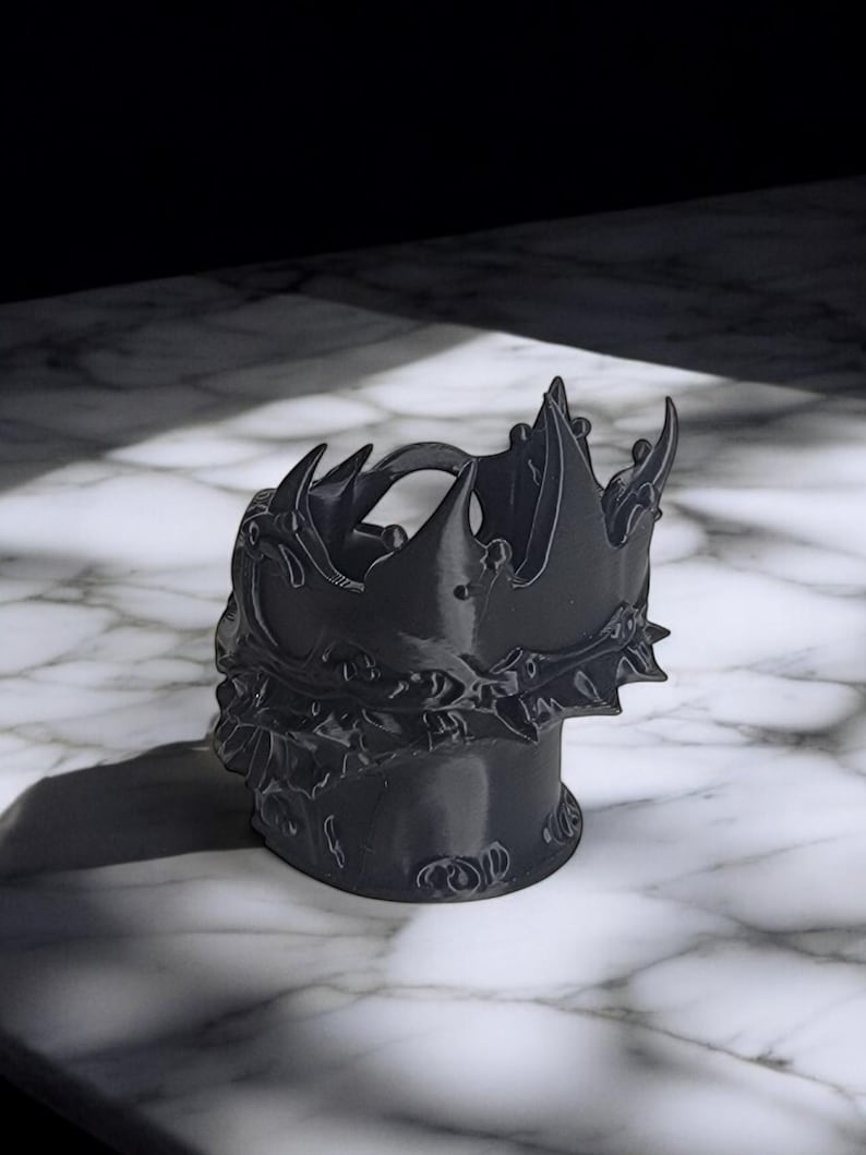 K&ouml;nnte beinhalten: Ein schwarzer, kronenf&ouml;rmiger Beh&auml;lter. Der Beh&auml;lter hat ein detailliertes Design mit scharfen Spitzen und eine strukturierte Oberfl&auml;che. Er steht auf einer Marmoroberfl&auml;che.