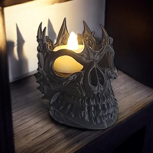 K&ouml;nnte beinhalten: Ein grauer, sch&auml;delformiger Kerzenhalter mit Kronen-Design. Er enth&auml;lt eine flackernde LED-Kerze, die Schatten auf das Holzregal wirft. Der Sch&auml;del hat detaillierte Merkmale und eine gotische &Auml;sthetik.