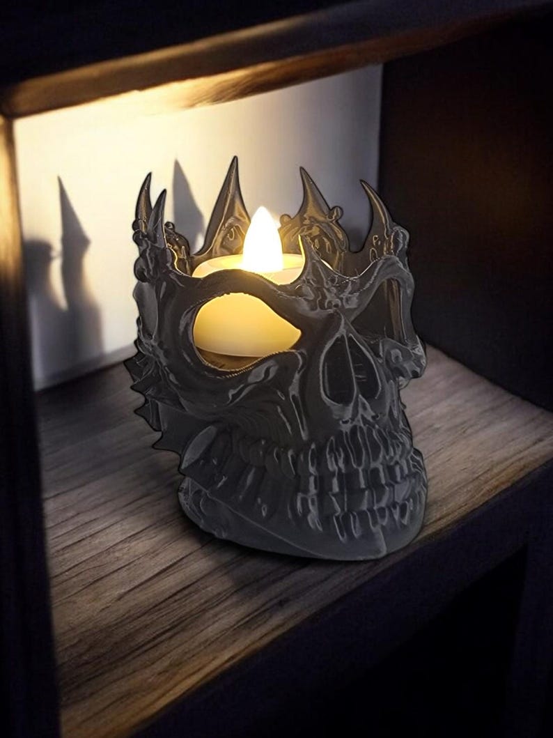 K&ouml;nnte beinhalten: Ein grauer, sch&auml;delformiger Kerzenhalter mit Kronen-Design. Er enth&auml;lt eine flackernde LED-Kerze, die Schatten auf das Holzregal wirft. Der Sch&auml;del hat detaillierte Merkmale und eine gotische &Auml;sthetik.