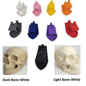 K&ouml;nnte beinhalten: Eine Auswahl an 3D-gedruckten Bootsmodellen in verschiedenen Farben, darunter Wei&szlig;, Rosa, Gelb, Orange, Grau, Blau, Lila und Rot. Das Bild zeigt auch zwei Sch&auml;delmodelle in dunklem und hellem Knochenwei&szlig; sowie ein schwarzes Bootsmodell.