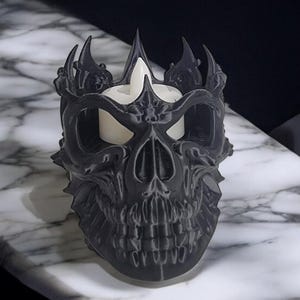 K&ouml;nnte beinhalten: Ein schwarzer, sch&auml;delartiger Kerzenhalter mit Kronenmuster. Der Kerzenhalter hat eine detaillierte, strukturierte Oberfl&auml;che und h&auml;lt eine wei&szlig;e Kerze. Das Design ist gotisch und dekorativ, passend f&uuml;r Halloween oder Themen-Dekoration.