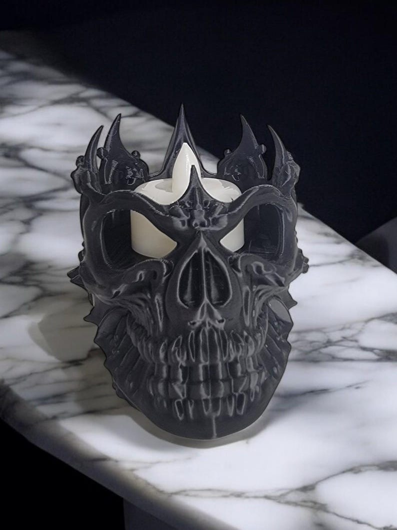 K&ouml;nnte beinhalten: Ein schwarzer, sch&auml;delartiger Kerzenhalter mit Kronenmuster. Der Kerzenhalter hat eine detaillierte, strukturierte Oberfl&auml;che und h&auml;lt eine wei&szlig;e Kerze. Das Design ist gotisch und dekorativ, passend f&uuml;r Halloween oder Themen-Dekoration.