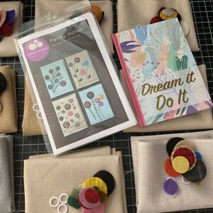 May include: A craft kit for making small fabric notebooks. The kit includes a pattern for four different designs, fabric scraps in various colors, and a small notebook with the text "Dream it Do it" on the cover.