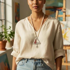 May include: A woman wearing a cream-colored linen shirt and light blue jeans. She is wearing a long pearl necklace with a silver pendant. The pendant is a triangle shape with a red and silver design.