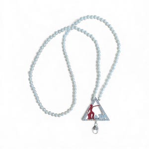 May include: A pearl beaded lanyard with a triangular pendant. The pendant features a red silhouette of a person, a Greek letter design, and is encrusted with small, clear crystals. A silver-colored clasp is attached.