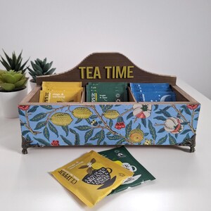 May include: A wooden tea bag organiser with a blue floral pattern, including lemons and flowers. The words "TEA TIME" are displayed on top. The organiser has three compartments filled with tea bags. Decorative metal feet complete the design.