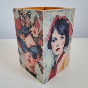 Puede incluir: Un portalápices cuadrado de madera con un diseño de collage. Un lado muestra el retrato de una persona con un pañuelo rojo, mientras que el lado adyacente muestra a una persona con alas de mariposa azules sobre los ojos entre flores.