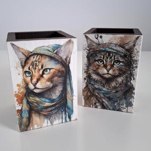 May include: Two decorative wooden boxes with watercolor cat portraits. Each box features a different cat wearing a blue scarf and head covering. The boxes have dark brown tops and are set against a white background.