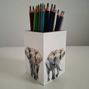Może przedstawiać: Biały drewniany pojemnik na ołówki z dwiema ilustracjami słoni. Pojemnik jest wypełniony różnymi kredkami. Kredki są ustawione pionowo, gotowe do użycia. Ilustracje słoni są szczegółowe i realistyczne.
