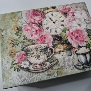 May include: Decorative box with a vintage tea party design. It features a teacup and saucer, a pocket watch, pink flowers, and a small silver cup. The background has a faded, antique aesthetic.