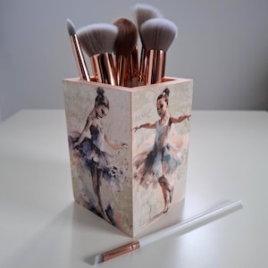 Puede incluir: Un portalápices de madera para brochas de maquillaje con una pintura de bailarina en los lados, lleno de varias brochas de maquillaje. El portalápices es rectangular y tiene un fondo beige claro. Las brochas tienen mangos de oro rosa y cerdas suaves.