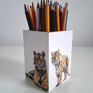 Puede incluir: Un portalápices de madera blanco con una ilustración de acuarela de dos tigres. El portalápices está lleno de una variedad de lápices de colores. Los lápices son de varios colores, incluyendo amarillo, naranja, marrón y azul.