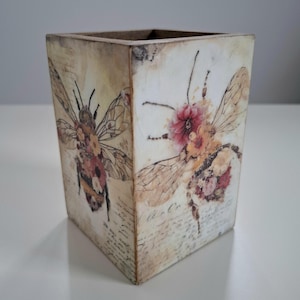 May include: A square wooden box with a distressed finish, featuring bee illustrations adorned with floral elements. The design incorporates vintage script and a warm colour palette, creating a rustic, decorative piece.