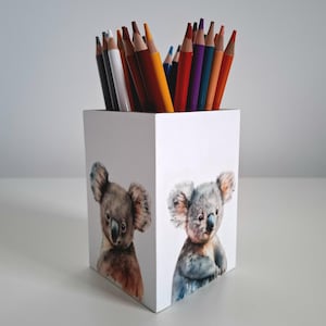 Puede incluir: Portalápices cuadrado blanco con ilustraciones de osos koala en acuarela. El portalápices está lleno de una variedad de lápices de colores en tonos rojos, amarillos, morados y negros. Un accesorio de escritorio decorativo.