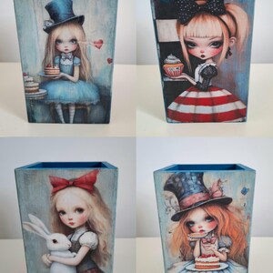 May include: A set of four square wooden pencil holders featuring illustrations of characters with large eyes. The designs include a figure in a blue top hat with cake, a figure with a cupcake labeled EAT ME, a figure holding a white rabbit, and a figure in a hat with cake.