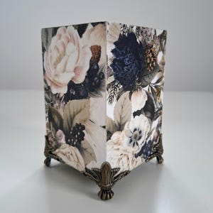 Puede incluir: Una caja decorativa cuadrada con un estampado floral con grandes flores de color rosa claro y azul oscuro, y hojas verdes. La caja tiene patas ornamentadas de color bronce.