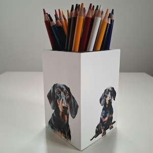 Puede incluir: Un portalápices de madera blanco con dos ilustraciones de perros salchicha. El portalápices está lleno de una variedad de lápices de colores. Los perros salchicha son negros y marrones con rasgos faciales detallados.