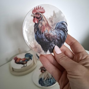 May include: A set of four round ceramic coasters featuring a watercolour illustration of a rooster. The coasters are white with a light brown border and are perfect for protecting surfaces from heat and spills.