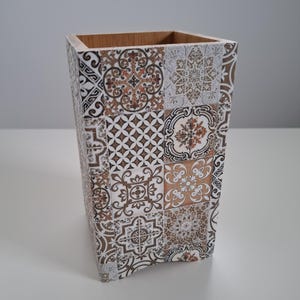 Puede incluir: Un recipiente decorativo de madera con forma cuadrada y diseño de mosaico. Los azulejos presentan una mezcla de patrones marrones, blancos y beige. El interior del recipiente es de color marrón claro.