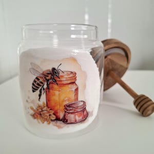 Puede incluir: Un tarro de vidrio transparente lleno de una sustancia blanca, con una ilustración de acuarela de una abeja y tarros de miel. Un cucharón de miel de madera y una tapa descansan junto al tarro. El tarro es probablemente para almacenar miel.