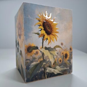 Puede incluir: Un bloque de madera con un diseño de campo de girasoles. El bloque muestra un gran girasol detallado en plena floración, con pétalos amarillos y un centro marrón oscuro. El fondo representa un cielo soleado y más girasoles.
