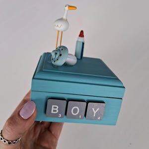 Puede incluir: Una caja de madera turquesa con la palabra "BOY" escrita con fichas de Scrabble. La caja está decorada con un chupete, un biberón y una figura de cigüeña. La caja está sostenida por una persona.