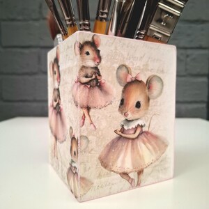 Puede incluir: Un portalápices cuadrado de madera decorado con imágenes de ratones con tutús de ballet rosas. El portalápices está lleno de pinceles. El fondo del portalápices tiene un aspecto vintage y descolorido.