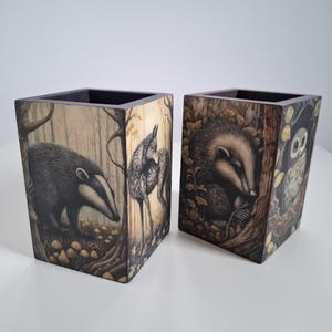 May include: Two wooden pen holders with forest animal illustrations. One features a badger and a bird in a forest scene, the other a badger and an owl. The pen holders are square with a dark interior.