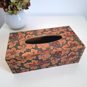 May include: A rectangular tissue box with a floral pattern in shades of orange, red, and blue. The box has an oval opening on top for tissue dispensing. The design features stylized flowers and leaves.