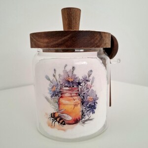 Könnte beinhalten: Ein klares Glas mit einem Holzdeckel und Griff, gefüllt mit einer weißen Substanz. Das Glas zeigt ein Aquarelldesign eines Honigglases, einer Biene und lila Blumen. Ein kleiner Holzlöffel liegt an der Seite.