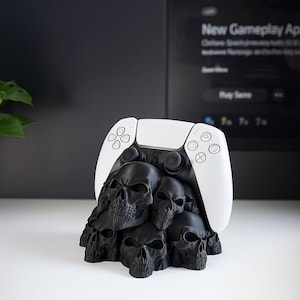May include: A white and black PlayStation 5 controller rests atop a black skull-shaped stand. The stand is composed of multiple detailed skulls, creating a unique and gothic aesthetic. The controller's buttons and joysticks are visible.