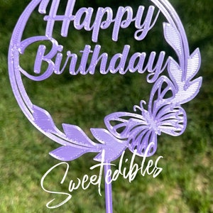 May include: Purple cake topper with a circle design that says "Happy Birthday" with a butterfly and leaves. The topper is on a stick and says "Sweetedibles" at the bottom.