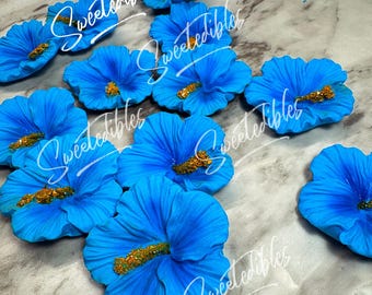 May include: Vibrant blue hibiscus flowers with gold centres are arranged on a marble surface. The flowers feature detailed petals and a textured look. The word "Sweetedibles" is written across the image in a cursive font.