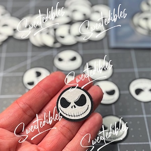 May include: A close-up of a hand holding a black and white Jack Skellington face edible image. The image is round and has a white background with black details. The image is surrounded by other Jack Skellington face edible images.