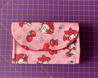 Puede incluir: Una cartera de tela rosa con cierre de botón rojo. La cartera tiene un patrón repetido de fresas y un personaje de dibujos animados con un lazo rojo.