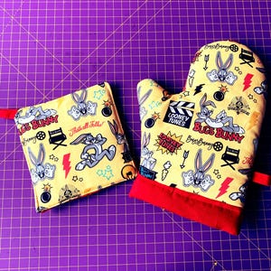 Könnte beinhalten: Zwei gelbe Ofenhandschuhe mit Bugs Bunny-Motiv. Die Handschuhe sind mit Bildern von Bugs Bunny, dem Looney Tunes-Logo und anderen Cartoon-Elementen verziert. Die Texte "Bugs Bunny" und "That's all folks!" sind auf den Handschuhen aufgedruckt.