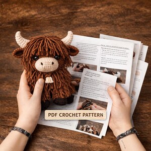 A crocheted Highland cow plushie with brown fur, cream-colored horns, and a cream-colored muzzle. The cow is held in front of a printed PDF crochet pattern. The text "PDF CROCHET PATTERN" is visible on the pattern.