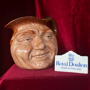 May include: A brown ceramic pitcher with a smiling face design. The pitcher has a handle and a spout. A white tag with blue text reads "Royal Doulton MADE IN ENGLAND".