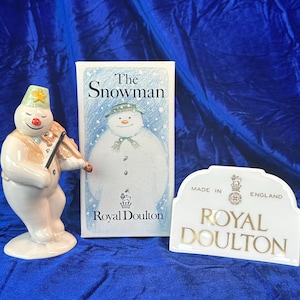 May include: A ceramic snowman figurine playing a violin, beside a book titled "The Snowman" and a white Royal Doulton sign. The snowman has a green hat with a yellow star, a red nose, and a tan vest. The book features a snowman illustration.