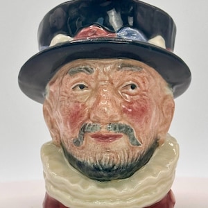 May include: A ceramic head figurine of a man wearing a black top hat with red, white, and blue details. The man has a mustache and beard, a ruffled collar, and a red base. This is likely a decorative item or collectable.