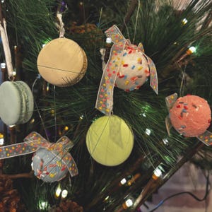 May include: Christmas tree ornaments in the shape of macarons and round balls. The ornaments are in various colors, including beige, light green, and pink, and are decorated with sprinkles and ribbons. The ornaments are hanging on a green Christmas tree.