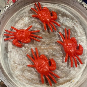 Puede incluir: Cuatro cangrejos de juguete de plástico rojo brillante sobre una superficie de madera blanca. Los cangrejos tienen ocho patas y una concha detallada.