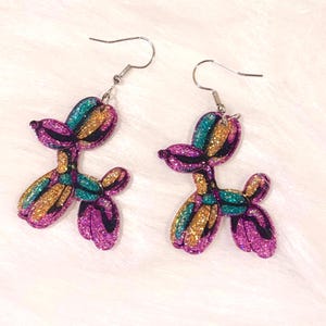 Könnte beinhalten: Ein Paar glitzernde, hundeförmige Ohrringe. Die Ohrringe haben ein farbenfrohes Design mit türkis, gold und lila Glitzer. Die Ohrringe haben silberfarbene Fischhaken-Ohrdrähte.