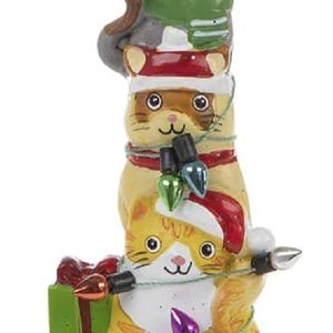 May include: A whimsical Christmas ornament featuring a stack of three cat figurines. The cats are adorned with festive hats, scarves, and string lights. The base of the ornament spells "meow" in green glitter.