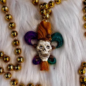 May include: A Mardi Gras necklace with gold beads and a skull pendant. The pendant features a skull with a jester's hat and colorful accents of purple, green, and orange. The beads are a shiny gold color.