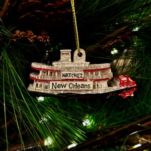 May include: A decorative Christmas tree ornament shaped like a steamboat. The ornament is white with red accents and features the words "Natchez" and "New Orleans". It hangs from a gold string against a backdrop of green pine needles.